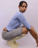 A woman crouching in a studio setting, wearing a light blue long-sleeve crop top, a gray skirt, and pale yellow socks against a solid light lavender background.
